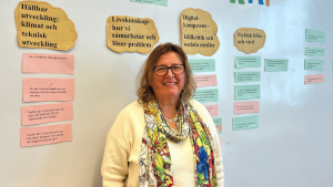 En kvinna med glasögon står framför en vägg och ler in mot kameran. På väggen finns lappar med text, så som "Livskunskap", "Digital kompetens" och "Psykisk hälsa och vård".