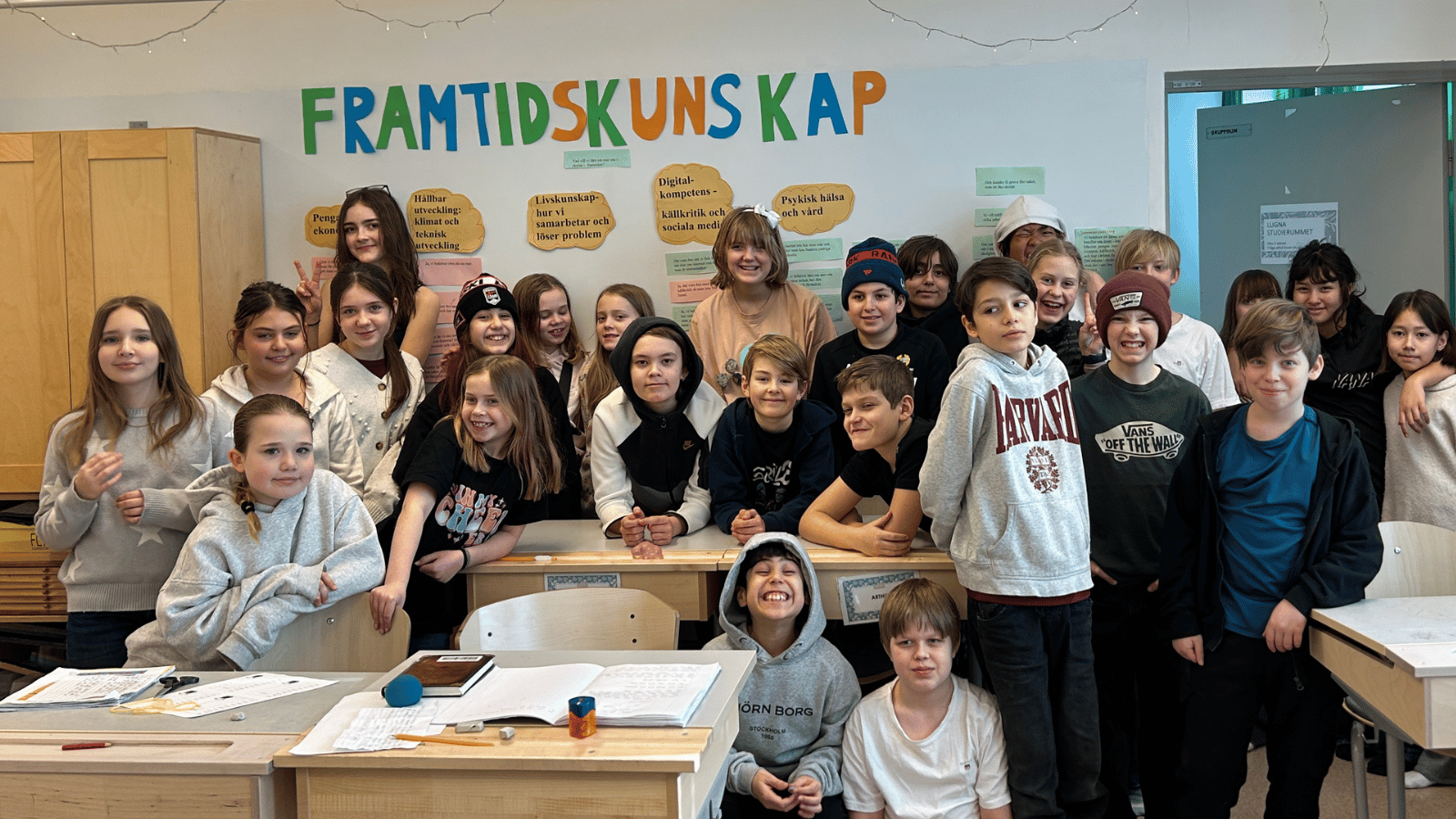 En mellanstadieklass står samlade i ett klassrum och tittar in i kameran. På väggen bakom dem står "FRAMTIDSKUNSKAP" med stora bokstäver.