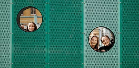 Tre tjejer kikar igenom två hål i en grön vägg.