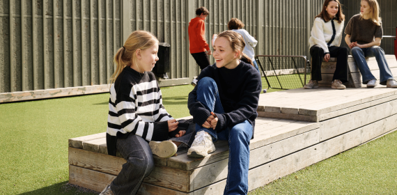 Två tjejer i mellanstadieåldern sitter på en träbänk på en skolgård och pratar.