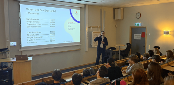 En man står längst fram i ett klassrum framför högstadieelever. Bredvid syns en skärm med text om vanliga yrken och dess löner.